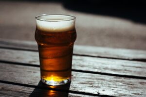 A pint of ale sits on top of a wooden bench, sun glistens through the glass and ale causing an amber reflection on the wood.